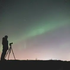 Sylwetka fotografa z aparatem na statywie pod rozgwieżdżonym niebem i zorzą polarną. Gwiazdy tworzą piękny efekt bokeh.