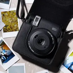 Czarny aparat Instax w etui, otoczony natychmiastowymi zdjęciami. Dowiedz się, jak robić zdjęcia Instax, tworząc własne, unikalne wspomnienia.