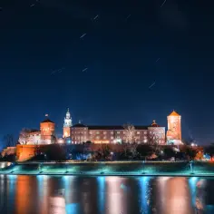 Nocne fotografowanie Wawelu z Wisły. Zamek oświetlony, na niebie smugi gwiazd.